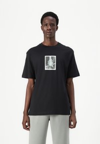Sort bomulds t-shirt med korte ærmer, der har en central monokrom grafisk illustration. Løs pasform og rund halsudskæring.