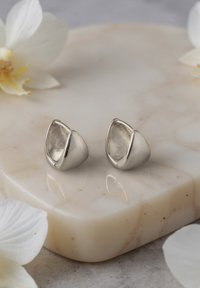 Orecchini d'argento con un design lucido e arrotondato e una leggera torsione. Esibiti su una superficie in marmo circondati da petali di fiori bianchi.