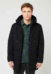Parka nera con cappuccio regolabile, chiusura con zip, tasche frontali e una camicia verde sottile al di sotto. Tessuto liscio, design informale.