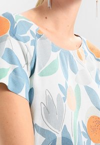 Floral blouse featuring a light background with blue, green, orange, and gray leaf patterns. Smooth texture with short sleeves.
