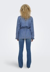 Veste en denim à taille cintrée, à manches longues et avec un col, vue de dos, assortie à un jean évasé de la même teinte bleue.