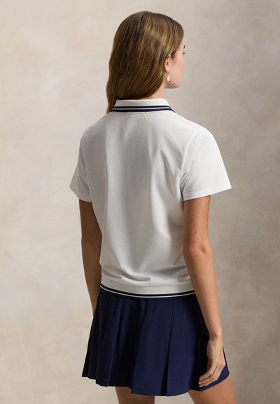 Λευκό πόλο μπλουζάκι με γιακά σε σκούρο μπλε αντίθεση, κοντά μανίκια, συνδυασμένο με φούστα σε σκούρο μπλε με πιέτες. Ομαλή υφή, καθημερινός σχεδιασμός, εφαρμοστή σιλουέτα.