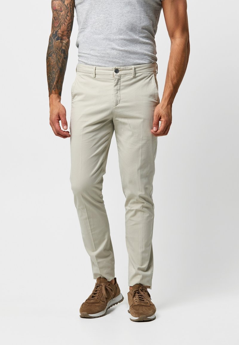 Chino chiari grigi con un taglio slim. Presentano una chiusura con bottone, tasche laterali e una texture liscia. Indossati con sneakers marroni.