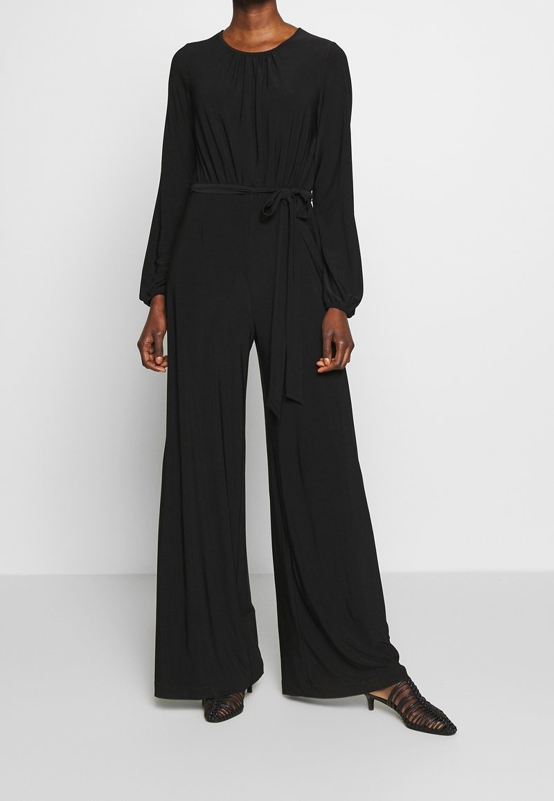 Femme portant une combinaison noire à manches longues avec des jambes larges et une ceinture nouée à la taille, assortie à des chaussures noires à brides et talons kitten.