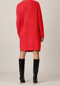 Robe tricotée rouge à manches longues, texture côtelée et coupe décontractée, associée à des bottes noires jusqu'aux genoux.