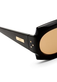 Μαύρος βραχίονας γυαλιών ηλίου με χρυσές λεπτομέρειες και φακό με κεχριμπαρένια απόχρωση, κοντινή λήψη στη μεντεσέ και στη λεπτομέρεια του λογότυπου στον βραχίονα.