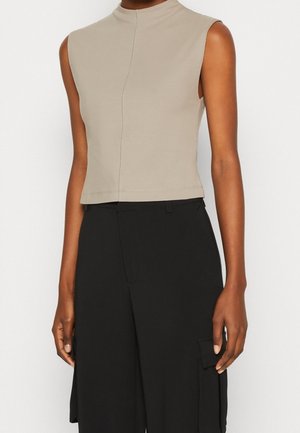 Beżowy top bez rękawów z wysokim kołnierzem, skrócony, zestawiony z czarnymi, szerokimi spodniami typu cargo z wysokim stanem na stojącej osobie.
