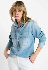 Lichtblauw denim overhemd met een knoopsluiting aan de voorkant, twee borstzakken en opgerolde mouwen. Heeft een vervaagde kleurverloop en zichtbare naden.