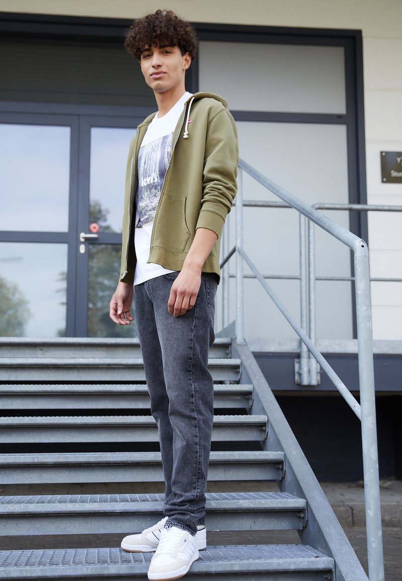 Oliven grønn zip-up hettegenser over en hvit grafisk T-skjorte, paret med mørk grå jeans og hvite sneakers, stående på metalltrapper.
