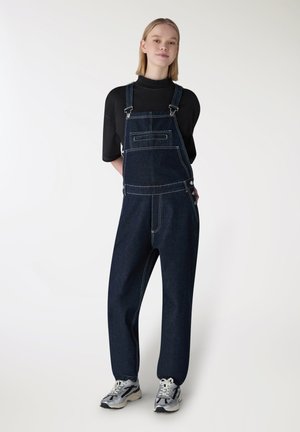 Giovane donna in piedi che indossa una salopette in denim scuro, una maglietta nera a maniche corte e sneakers grigio-bianco su sfondo bianco.