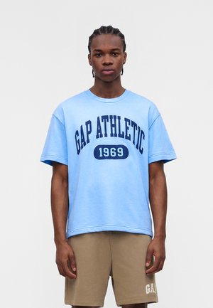 Νέος άνδρας με πλεκτά μαλλιά που φοράει ένα ανοιχτό μπλε κοντομάνικο μπλουζάκι "GAP ATHLETIC 1969" και μπεζ σορτς με το "GAP" στο αριστερό πόδι.