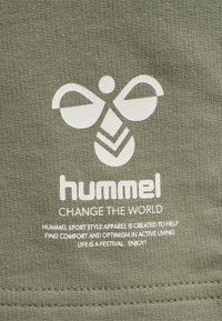 Textilie olivové barvy s hladkou strukturou má bílé potisknuté logo a text, který uvádí "hummel" a "CHANGE THE WORLD" v různých písmenech.
