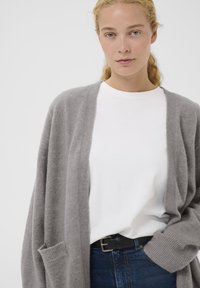 Cardigan lavorato a maglia grigio chiaro con apertura frontale, polsini a coste e tasche, abbinato a una t-shirt bianca e jeans in denim scuro.