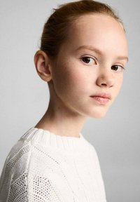 Nahaufnahme eines Person mit hellbraunen Haaren undSommerfreckles, die einen strukturierten weißen Pullover mit geripptem Halsausschnitt trägt, vor einem neutralen Hintergrund.