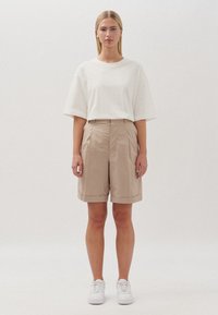 Pantaloni corti beige a vita alta con pieghe, abbinati a una t-shirt bianca morbida. Il modello indossa sneakers bianche. Design semplice e minimal.