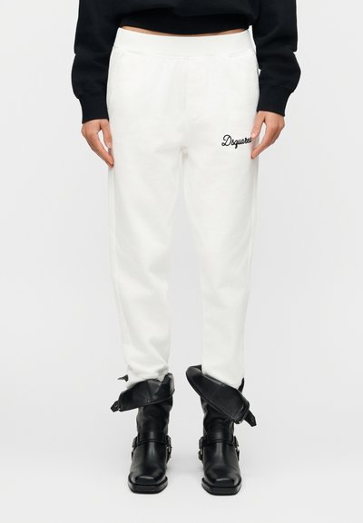 Un pantalon de survêtement blanc avec une ceinture élastique et des poches latérales, orné de lettres brodées noires sur la cuisse gauche. Porté avec des bottines noires.