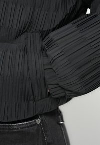 Μαύρο μπουφάν puffer με υφασμάτινη, πλισέ σχεδίαση. Διαθέτει ελαστική βάση και μανσέτες, με ομαλή επένδυση ορατή στο άνοιγμα του μανικιού.