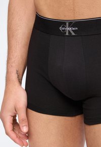 Man draagt zwarte Calvin Klein boxershorts, met zichtbaar logo op de tailleband en een gedeeltelijk zicht op de torso, arm en hand met een kleine tatoeage.