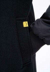 Ruka v černé polstrované rukavici sklouzne do kapsy černého vlněného kabátu s malou žlutou cedulkou zobrazující zavřené oko.