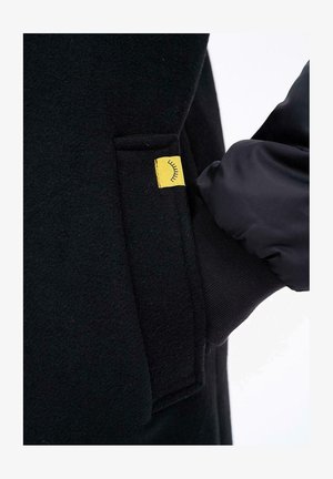 Ręka w czarnej, pikowanej rękawiczce wsuwa się do kieszeni czarnego wełnianego płaszcza z małą żółtą metką przedstawiającą symbol zamkniętego oka.