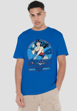 Man wearing blue t-shirt featuring comic-style Wonder Woman with lasso and “Wonder Woman” text on front, beige shorts, neutral background.