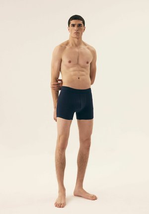 Marineblaue Boxer-Briefs aus glattem Stoff, mit einem weichen elastischen Bund. Das Modell steht barfuß mit sportlicher Figur vor einem neutralen Hintergrund.