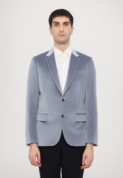 Blazer in velluto gradient blu con revers in satin, due tasche frontali e chiusura a un bottone. Indossato su una camicia bianca.