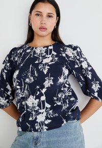 Navy Bluse mit weißem Blumenmuster, runder Ausschnitt und Puffärmeln. Strukturiertes Material, lockere Passform, kombiniert mit hellblauem Denim.