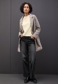 Cappotto in lana grigio chiaro sopra un maglione a girocollo giallo pallido e jeans neri a gamba larga, abbinati a stivaletti neri. Tessuto liscio, vestibilità su misura.