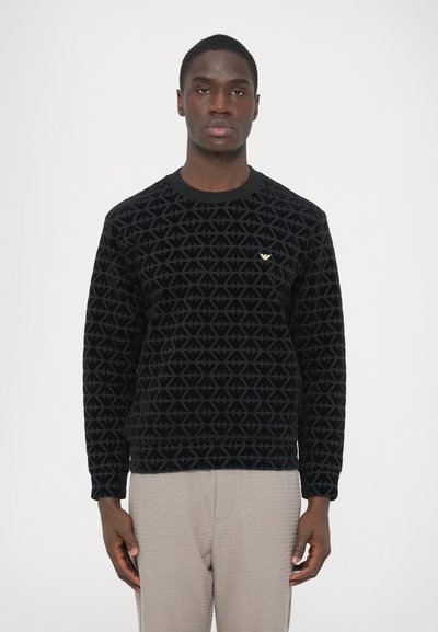 Black sweatshirt with a textured, geometric pattern, featuring a round neck and a small gold logo on the chest. Paired with light-colored pants.