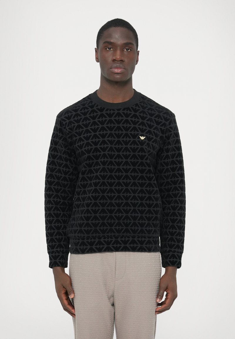 Čierna mikina s textúrovaným, geometrickým vzorom, s okrúhlym výstrihom a malým zlatým logom na hrudi. Kombinovaná so svetlými nohavicami.