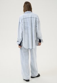 En ljusblå denimskjorta med rundad nederkant, knappmanchetter och detaljer vid baksidan, tillsammans med matchande löst sittande jeans och svarta skor.