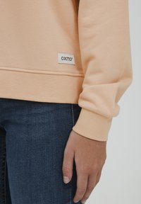 Felpa color pesca realizzata in morbido tessuto, con maniche a cuffia e un'etichetta logo Oxmo cucita vicino all'orlo. Indossata con jeans in denim scuro.