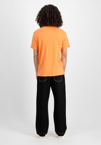Kurzes, oranges T-Shirt, das mit schwarzen, weit geschnittenen Hosen kombiniert ist, die weiße Nähte und einen geraden Saum haben. Die Schuhe sind aus Canvas mit einer dicken Sohle.