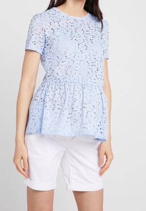 Kvinde iført lyseblå kortærmet blonderbluse med peplumskørt og hvide knælange shorts med opkrogede manchetter, stående foran en ensfarvet baggrund.