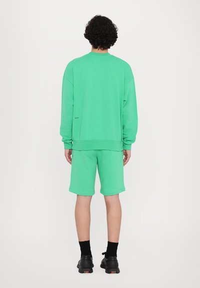 Pulover din bumbac verde, cu umeri lăsați, asortat cu pantaloni scurți matching. Ambele au o textură netedă și o croială relaxată. Pantofi negri și șosete.