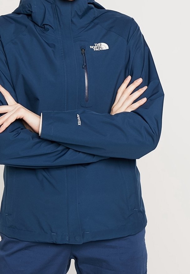 Personne portant une veste bleue The North Face GORE-TEX avec les bras croisés, montrant la fermeture éclair avant et la poche poitrine.