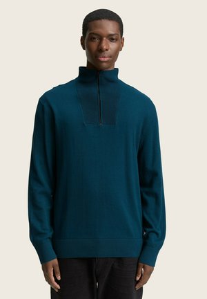 Pull-over en demi-fermeture éclair teal avec col et poignets côtelés. Texture lisse et design ajusté, associé à un pantalon sombre. Apparence simple et moderne.
