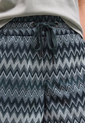 Detailní záběr černých a stříbrných kalhot se vzorem cikcak s přivázaným černým šňůrkem a částí světle šedé košile nahoře.