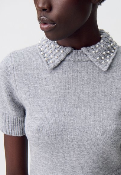 Suéter gris de punto con mangas cortas, que presenta un cuello texturizado adornado con pequeñas perlas blancas. Textura suave y fina en todo el suéter.