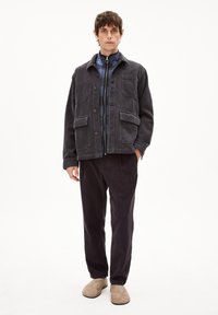 Dunkelgraue Cordjacke mit zwei vorderen Taschen, kombiniert mit einem navyblauen karierten Hemd und dunkelbraunen Cordhosen. Beigefarbene Schuhe.