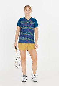Ung kvinde iført sportsbeklædning, der holder en badmintonracket, står og smiler mod en simpel hvid baggrund.