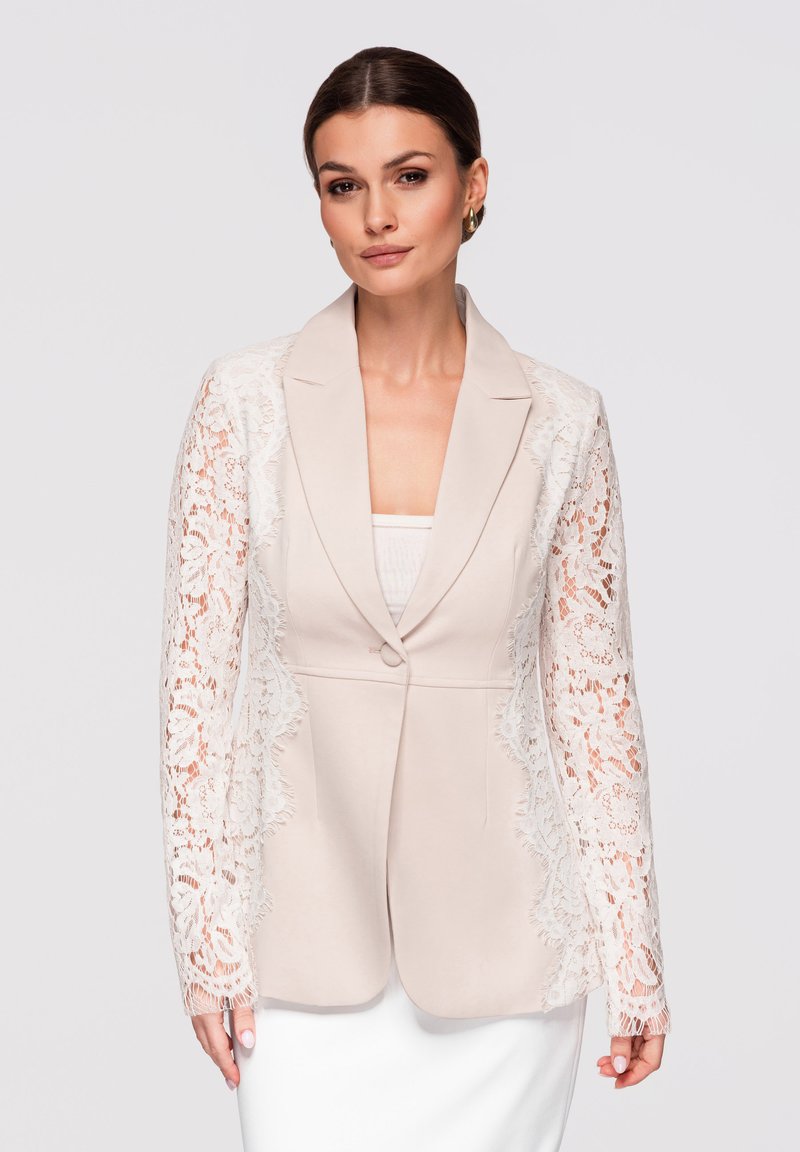 Femme portant un blazer beige avec des manches longues en dentelle et un seul bouton, associé à un haut blanc et une jupe, sur un fond uni.