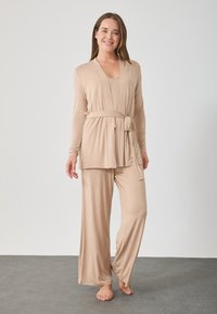 Beige σετ lounge wear με μακρυμάνικο τοπ wrap που δένει και παντελόνι με φαρδιά πόδια, φτιαγμένο από απαλό ύφασμα με άνετη εφαρμογή.