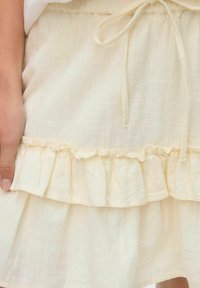 Jupe beige clair à volants fabriquée en tissu texturé. Dotée d'une taille élastique avec un lien et de deux couches de volants à l'ourlet.