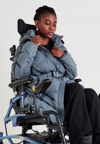 Pilkas pūkinis paltas su gobtuvu ir diržu, pasižymintis įstrižais siūlėmis ir vandeniui atsparia tekstūra, dėvimas asmens, esančio neįgaliojo vežimėlyje.