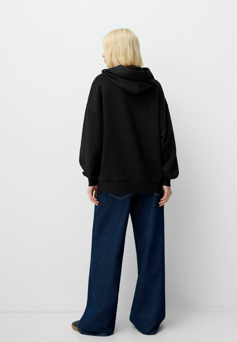 Felpa nera oversize con cappuccio regolabile, tessuto strutturato e maniche ampie, abbinata a jeans in denim blu a vita alta e gamba larga.