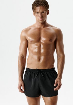 Muskułowy mężczyzna bez koszulki z mokrą skórą, ubrany w czarne kąpielówki ze sznurkiem, stojący na jednolitym białym tle.