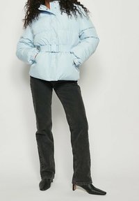 Lyseblå dunjakke med bælte i taljen, lange ærmer og elastiske manchetter; kombineret med sorte jeans og spidse sorte hæle.