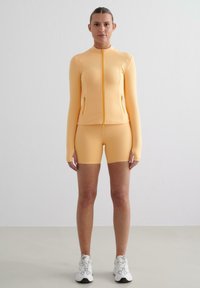ŽLTÝ športový outfit, ktorý obsahuje zipsovú dlhú rukávovú blúzku s bočnými vreckami a zodpovedajúce šortky. Hladká, elastická tkanina s priliehavým strihom.